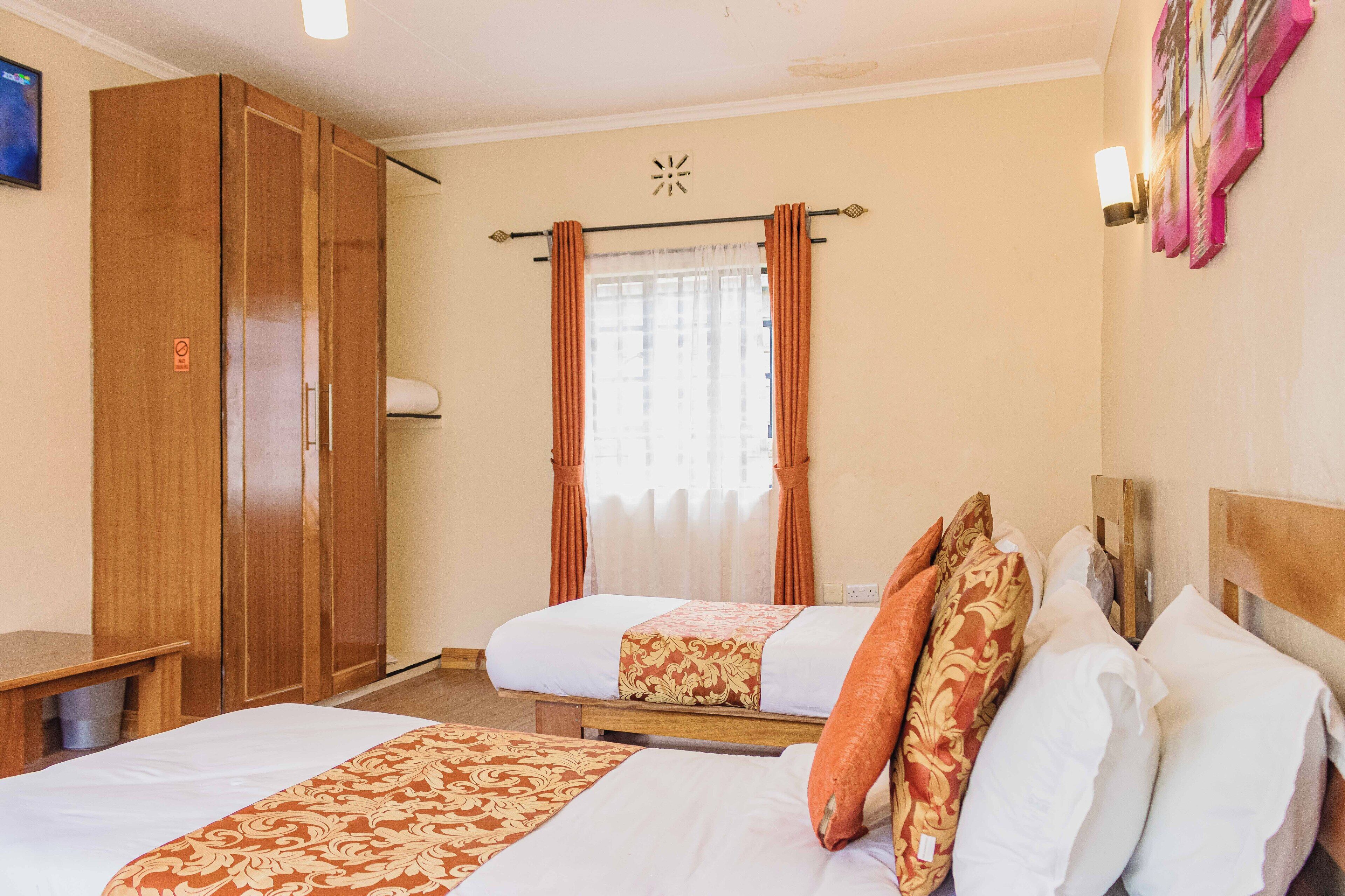 A hotel room with two beds, wooden wardrobe, curtains, and a wall-mounted light fixture.