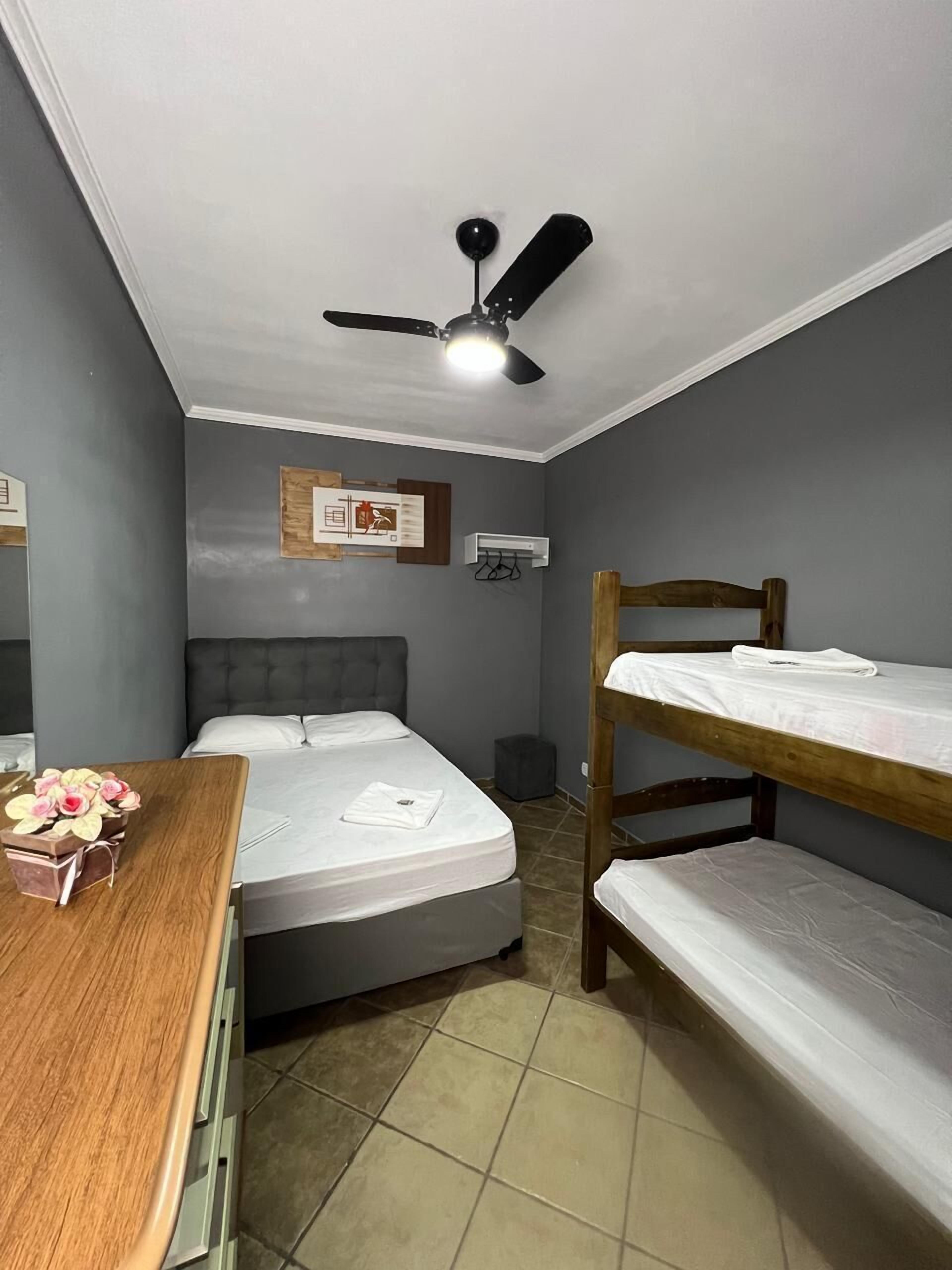 Una habitación con una cama individual, una cama nido de madera, un escritorio con un jarrón de flores, un ventilador de techo y un aire acondicionado de pared.