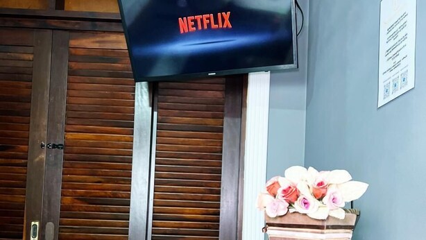 A room with a wooden door, a wall-mounted television displaying 'NETFLIX', and a remote control on a wooden table. - HOSTEL ANPRADO (Guarulhos)