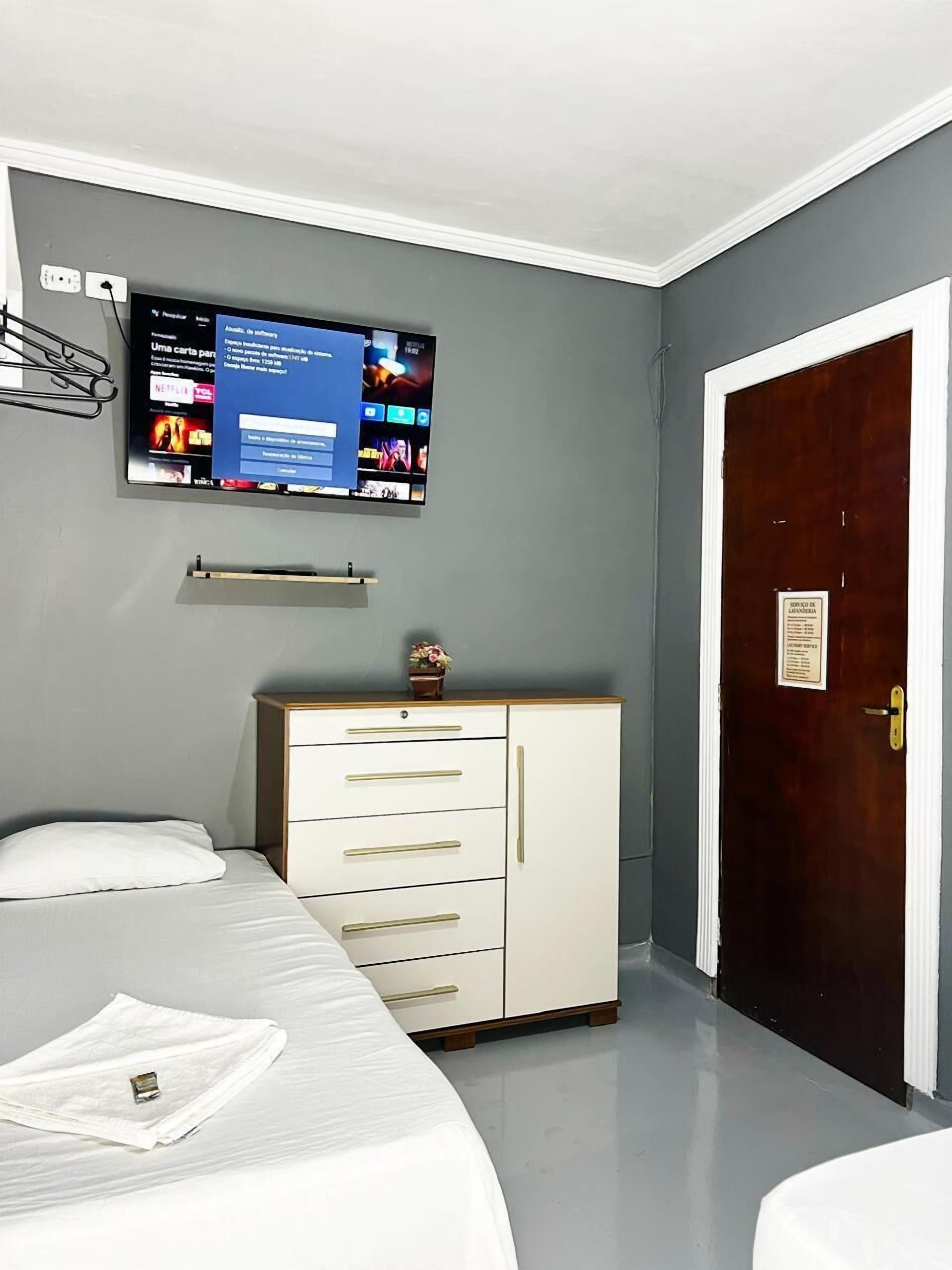 A hotel room with a bed, a dresser, a television mounted on the wall, and a door with a sign.