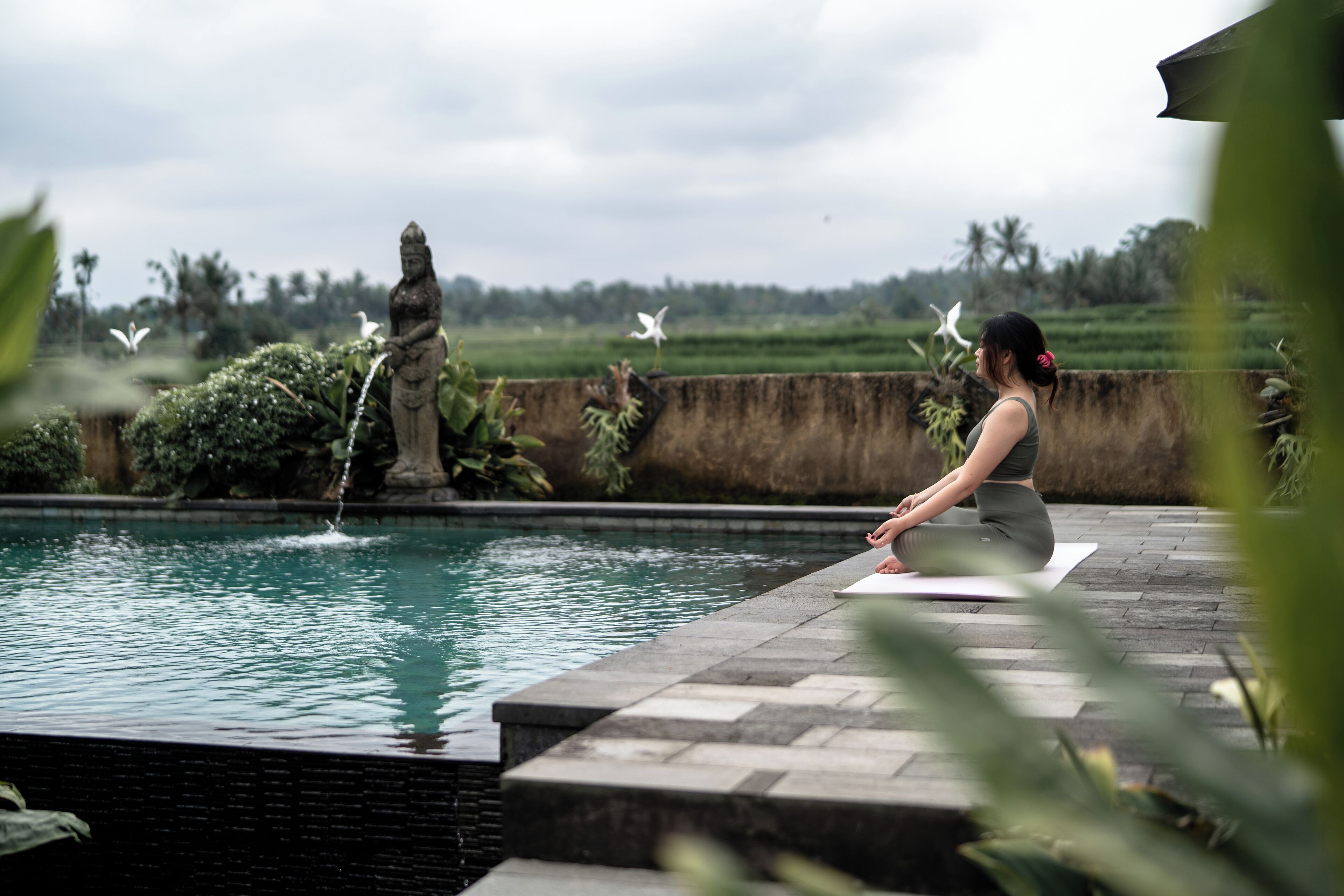 Une personne médite au bord d’une piscine, avec une statue et de la verdure en arrière-plan.