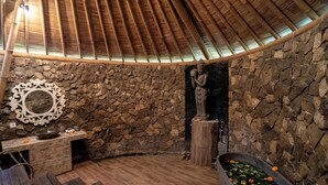 A stone-walled room with a wooden bench, a stone bathtub with flowers, and a statue. - Padi Bali Retreat (Payangan)