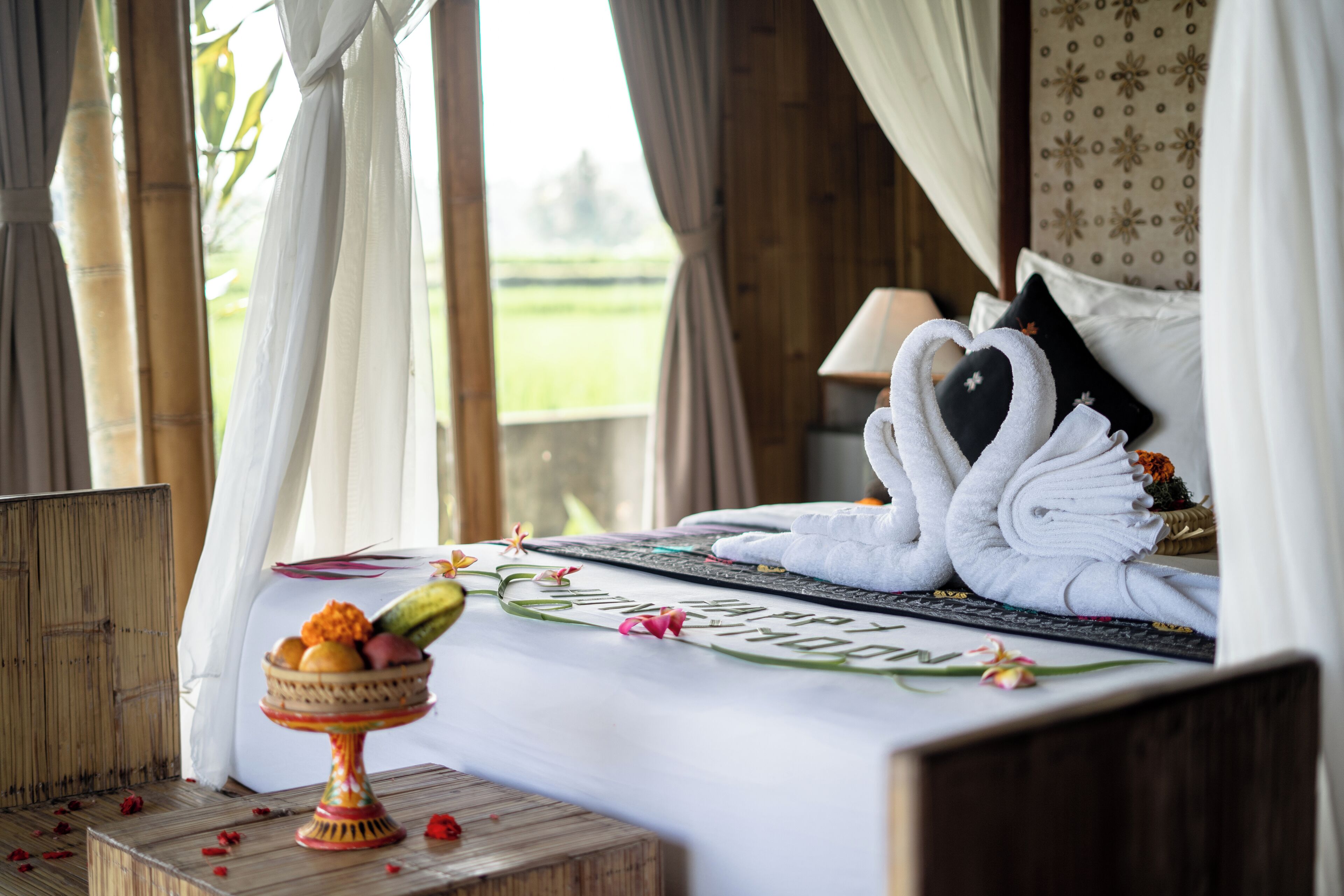 Una cama bien tendida con una decoración de toallas con forma de cisne, un frutero sobre una mesa y vistas a un paisaje verde a través de la ventana.
