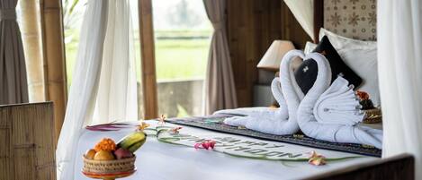 Una cama bien tendida con una decoración de toallas con forma de cisne, un frutero sobre una mesa y vistas a un paisaje verde a través de la ventana.