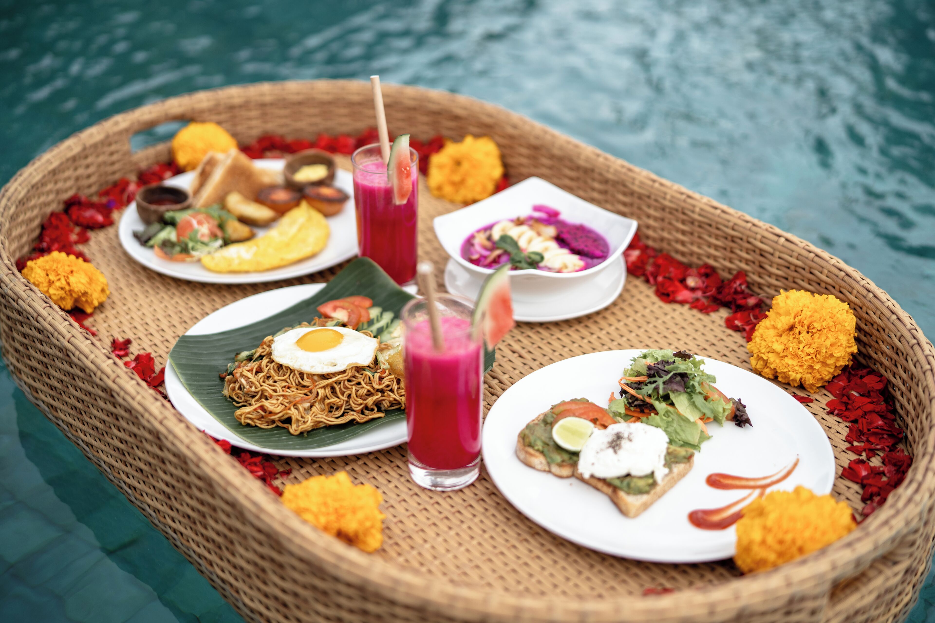 Bandeja de vime com diversos pratos, incluindo noodles, salada e um smoothie, colocada em ambiente de piscina.