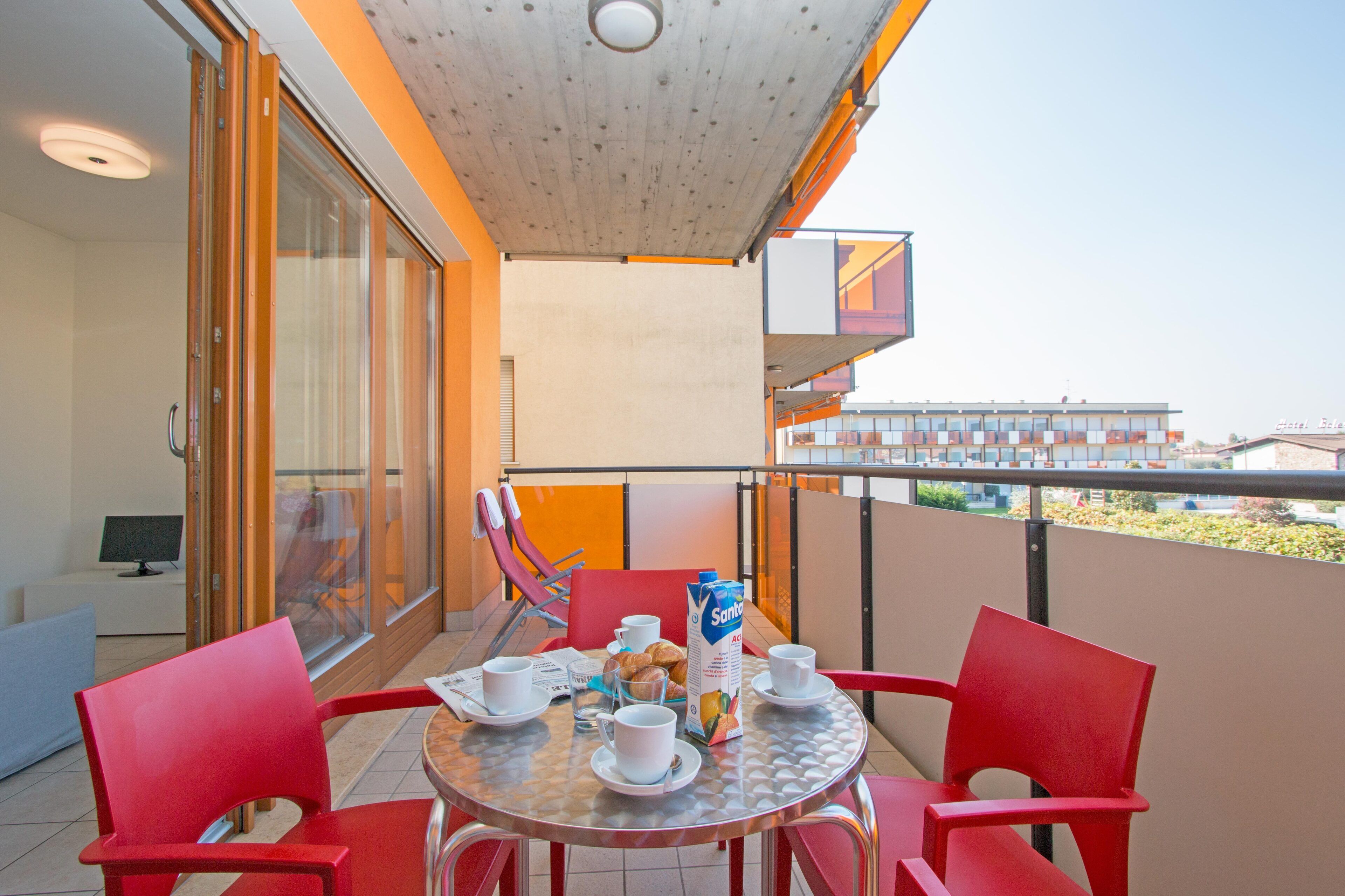 Un balcon avec une table dressée pour le petit-déjeuner, des chaises rouges et une porte vitrée donnant sur l’intérieur.
