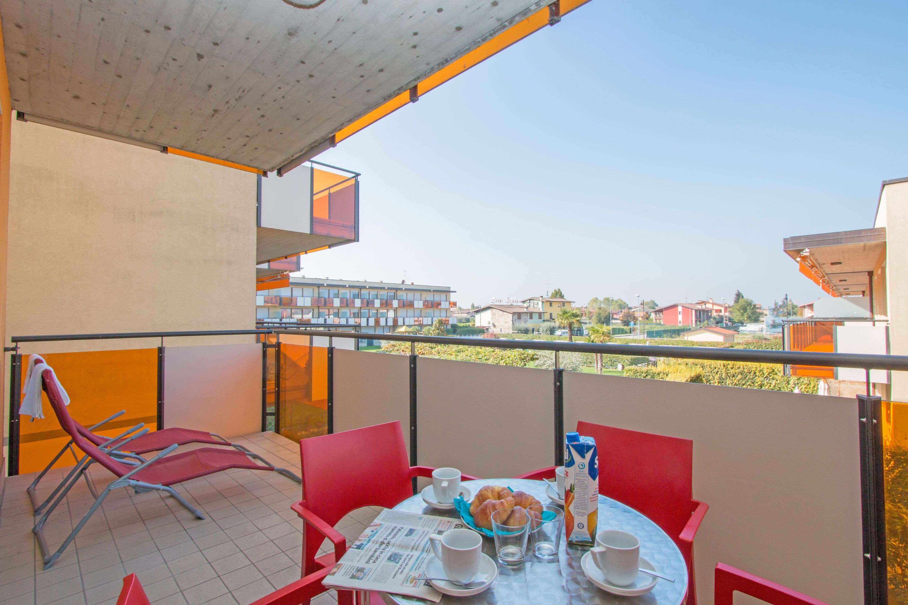 Un balcon avec des chaises rouges, une table où sont disposés des aliments pour le petit-déjeuner, et une vue sur des bâtiments au loin.
