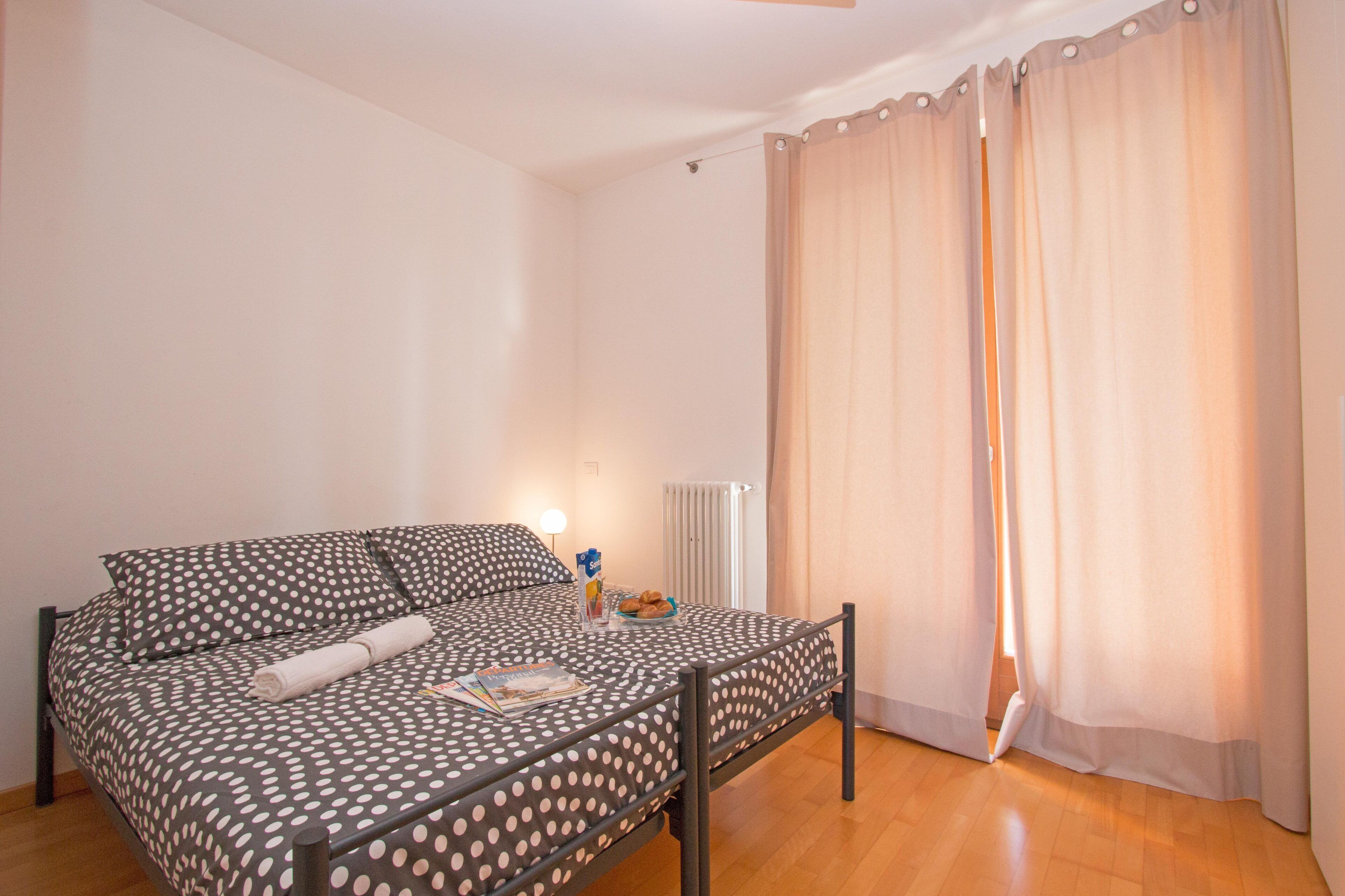 A single bed with a polka dot bedspread, a bedside table with a lamp, and a window with curtains.