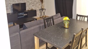 Un saló modern amb un sofà gris, una taula de dinar amb cadires, un televisió de pantalla plana i un ram de flors grogues decoratiu.