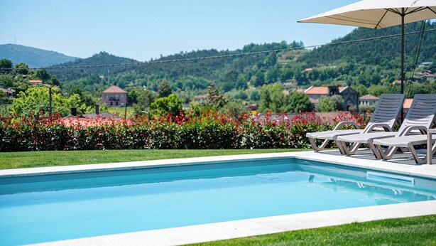 A swimming pool with lounge chairs and an umbrella, surrounded by greenery and houses in the background. - Brook House - T3 - Mosteiro, Vieira do Minho (Vieira do Minho)