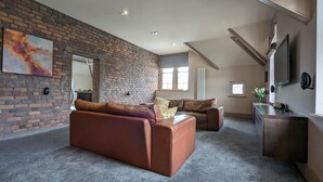 A spacious living room with a brick wall, a brown leather sofa, a second sofa, a large mirror, and a flat-screen TV mounted on the wall. - Penthouse-highstreet-sleeps2-87ws (Merthyr Tydfil)