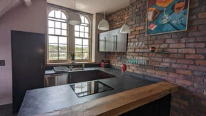 A modern kitchen with a large island, a window with arched top, and a brick wall with a framed artwork. - Penthouse-highstreet-sleeps2-87ws (Merthyr Tydfil)