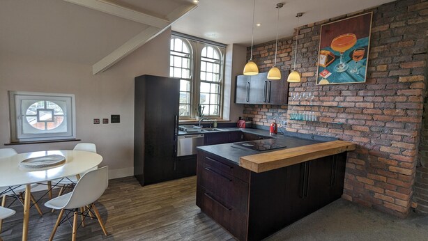 A modern kitchen with a central island, wooden flooring, and a brick accent wall. - Penthouse-highstreet-sleeps2-87ws (Merthyr Tydfil)
