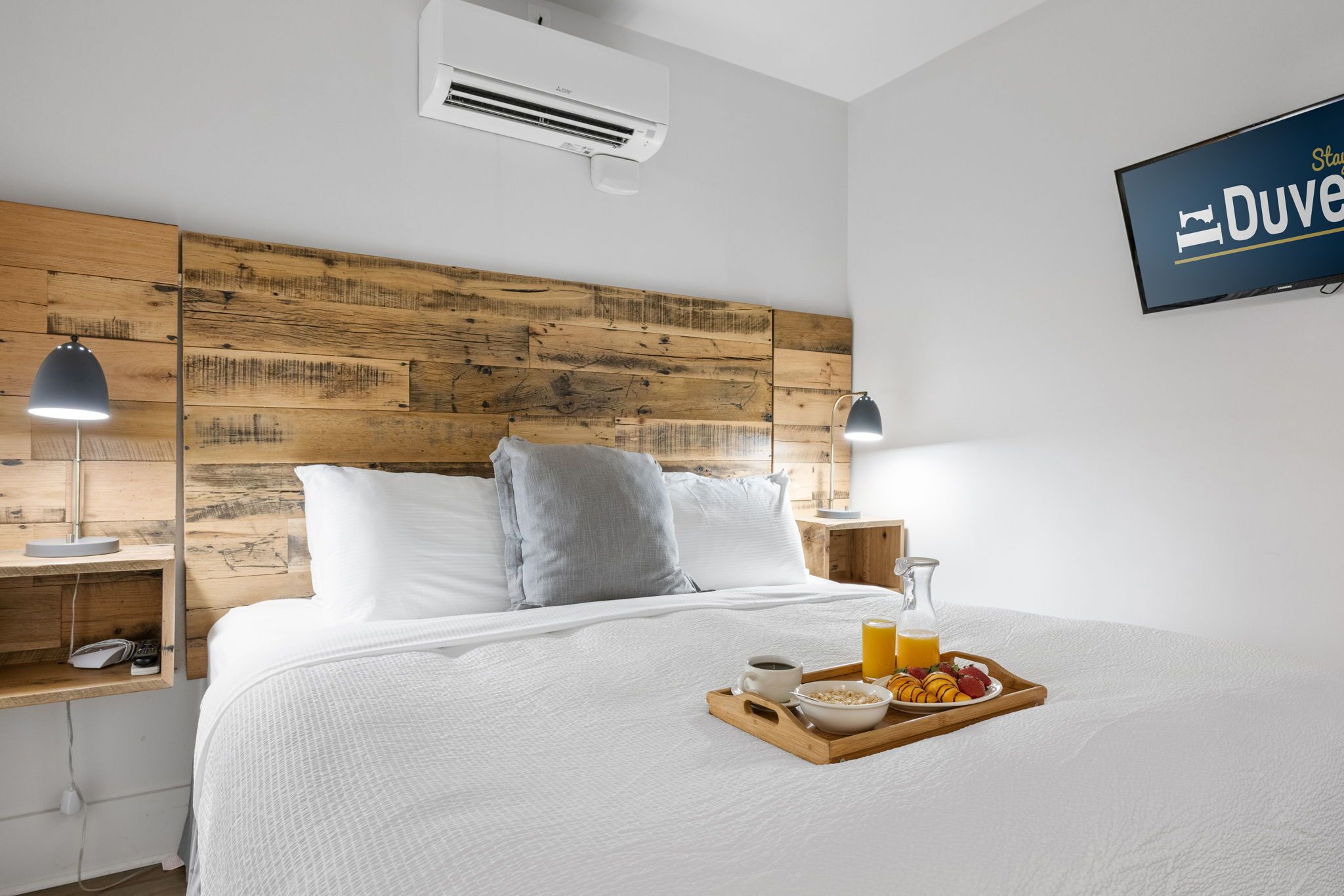 Et hotelværelse med en træsenggavl, en seng med hvide sengetæpper, sengeborde med lamper og et bakke med morgenmad, samt en aircondition.