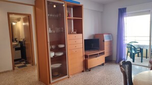 A compact living space with a kitchenette, a wooden cabinet with shelves, a television, and a dining area with plastic chairs. - Apartamento muy Fresco Verano (VC)