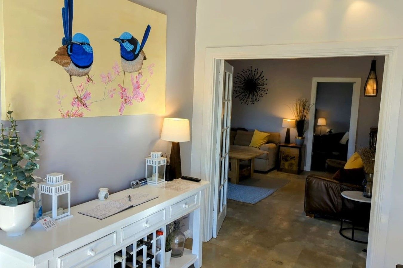Un salon avec une console blanche, un tableau représentant des oiseaux, une plante en pot et un canapé agrémenté de coussins jaunes.