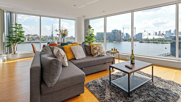 A modern living room with a large sofa, a coffee table, and a view of a city skyline and river. - MARINA VIEW HAVEN- Stylish 2-Bedroom Retreat Near Excel London. (London)