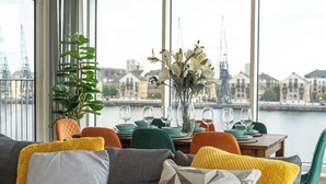 A dining area with a view of a river and buildings, featuring a table set with green plates, wine glasses, and a vase of flowers. - MARINA VIEW HAVEN- Stylish 2-Bedroom Retreat Near Excel London. (London)