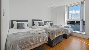 A modern bedroom with two beds, a large window with a view of a river and buildings, and a wooden floor. - MARINA VIEW HAVEN- Stylish 2-Bedroom Retreat Near Excel London. (London)