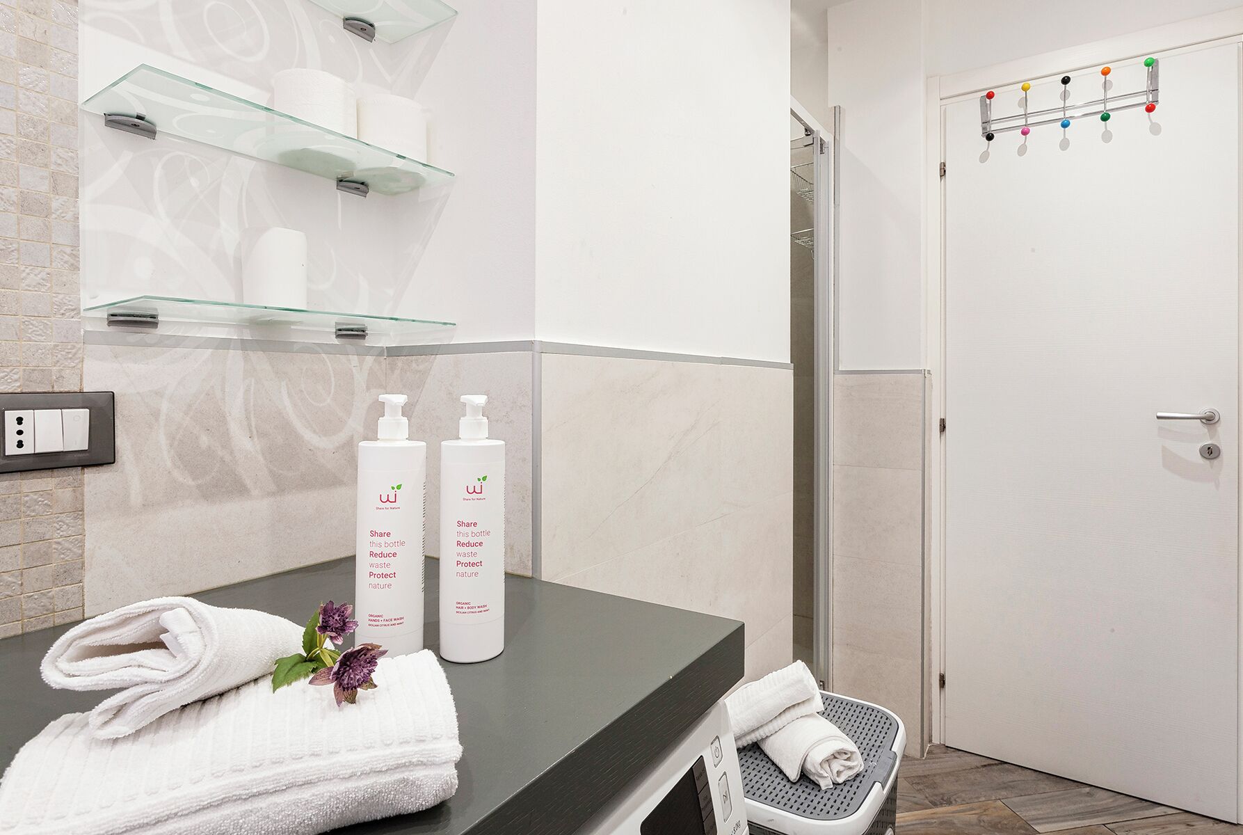 Une salle de bain avec une étagère en verre où sont rangés les articles de toilette, une serviette posée sur le comptoir et un espace douche.