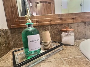 A bathroom counter with a green bottle labeled 'MOUTHWASH,' a stack of beige cups, and a glass jar with a white towel. - ✨ Seasons at BlueSky - Lux Penthouse 🏔️ Ski In/Ski Out 🎿 Spa 🧖‍♀️Resort Pool (Breckenridge)