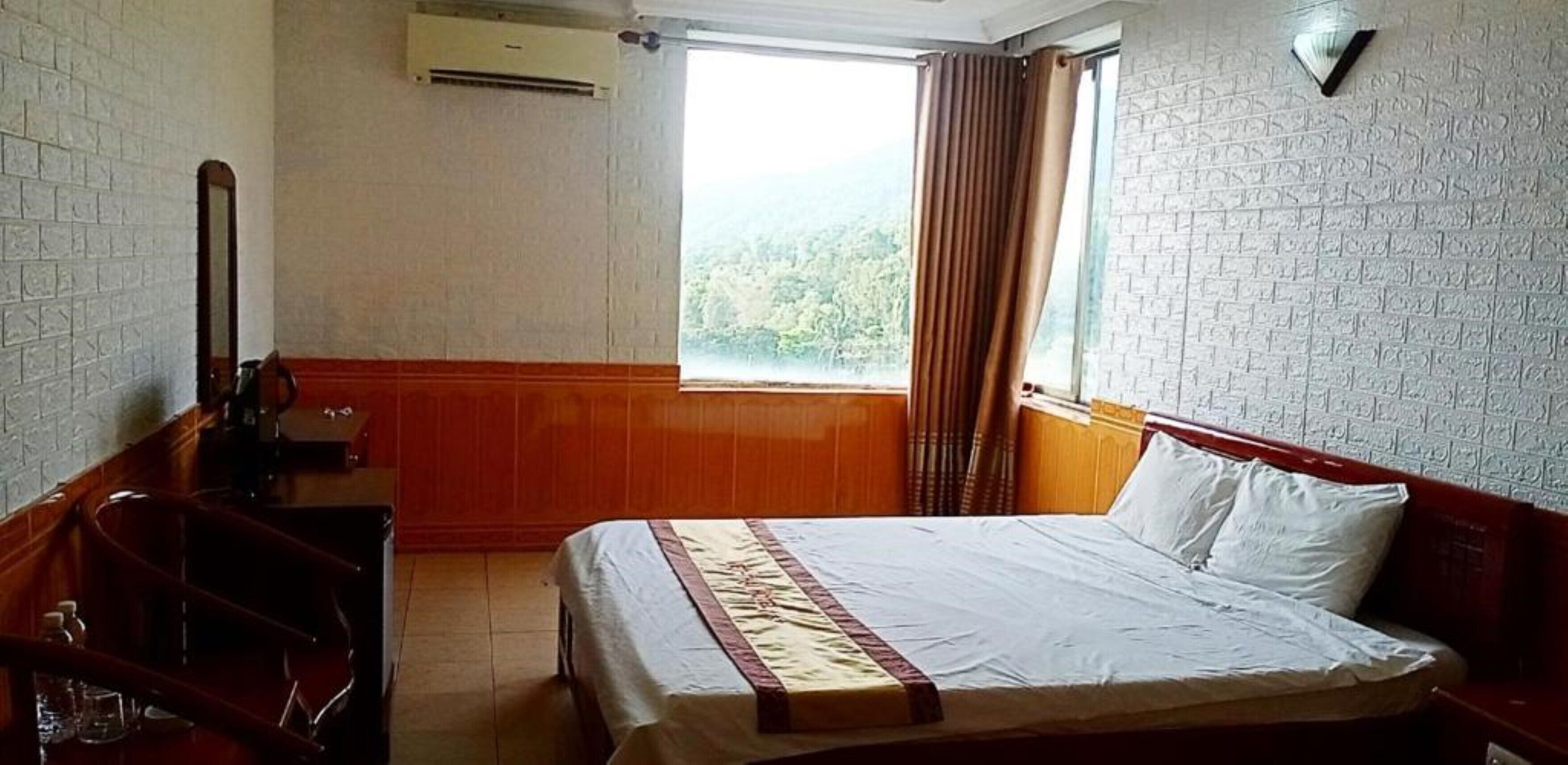 A hotel room with a bed, a wooden desk, a mirror, and a window with curtains.