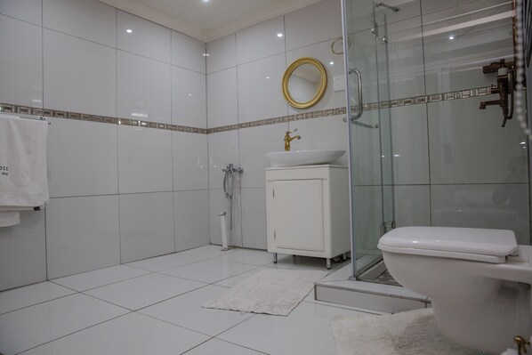 A modern bathroom with white tiles, a glass shower enclosure, a white sink with a gold faucet, and a white toilet. - Melrose Hideout B&B (Johannesburg)