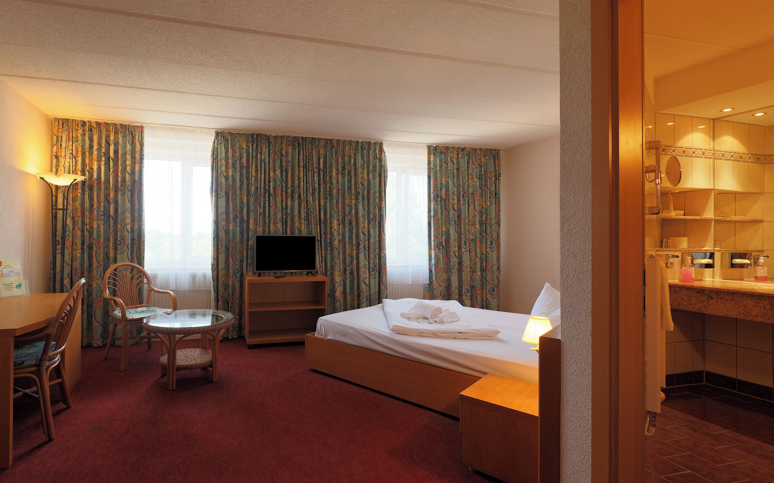 Habitación de hotel con cama, escritorio con silla, televisión y baño visible a través de una puerta abierta.