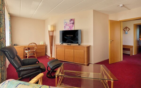 A hotel room with a flat-screen TV, a glass coffee table, two leather chairs, a wooden cabinet, and a red carpet. - AXXON Hotel (Brandenburg an der Havel)