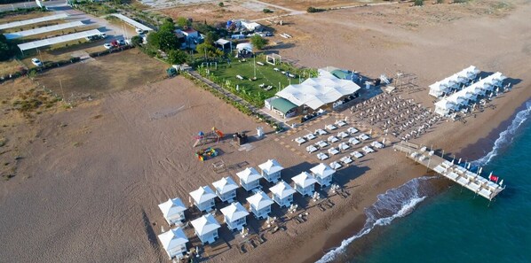 An aerial view of a beachside hotel with multiple white-roofed buildings, a large covered area, and beach umbrellas.