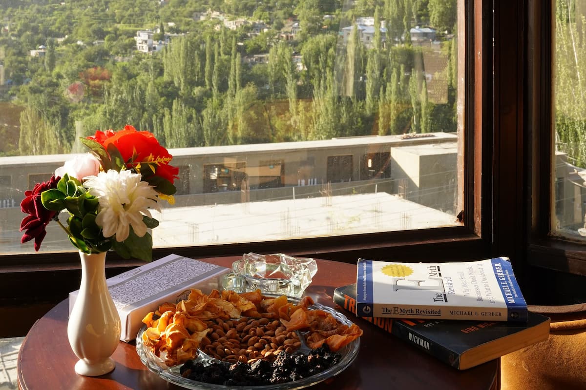 Ein Tisch mit einem Buch, einer Blumenvase und einer Schale Nüsse vor einem Fenster mit Blick auf eine Berglandschaft.