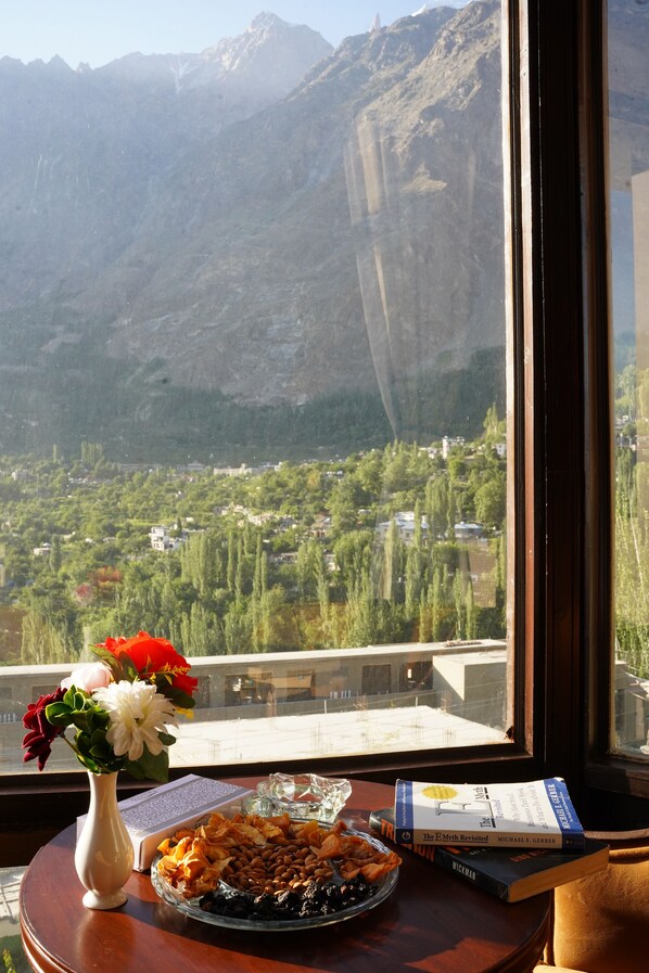 A table with a book, a vase of flowers, and a plate of nuts by a window overlooking a mountainous landscape. - Hunza Darbar Hotel  (Karimabad)