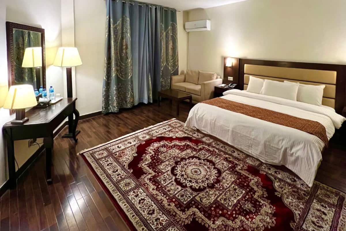 Ein Hotelzimmer mit einem Bett, einem Schreibtisch mit Lampen, einem Sessel und einem Deckenventilator, sowie einem großen, gemusterten Teppich.