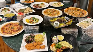 Una mesa de buffet con diversos platos, entre ellos pizzas, ensaladas y carnes, frente a grandes ventanales y jarrones decorativos.