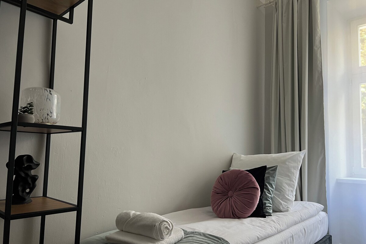 A neatly made bed with a grey blanket, a pink pillow, and a white pillow. There is a shelf with decorative items and a rolled white towel on the bed.