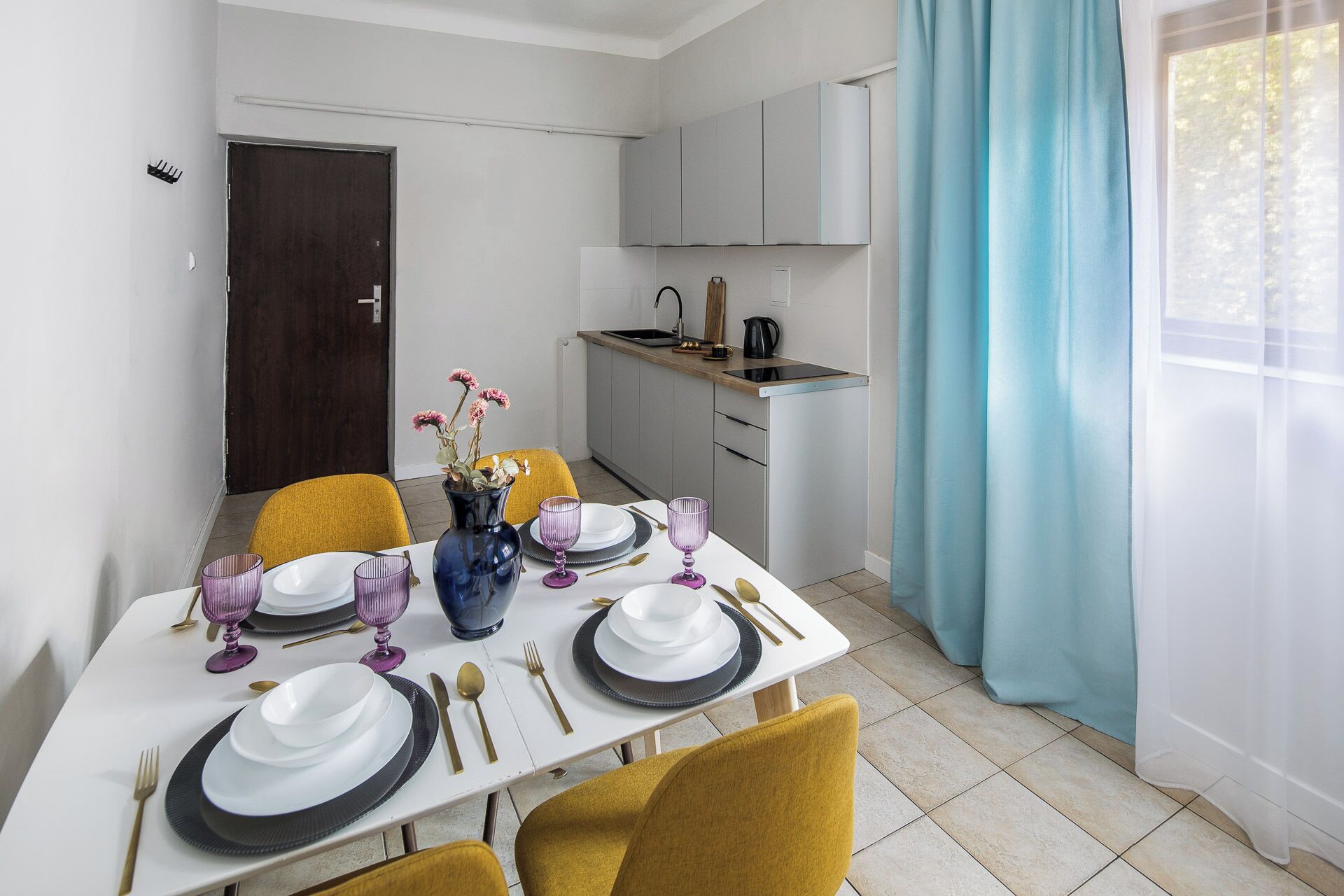 Un coin repas avec une table blanche dressée pour quatre personnes, des chaises jaunes et un vase rempli de fleurs.