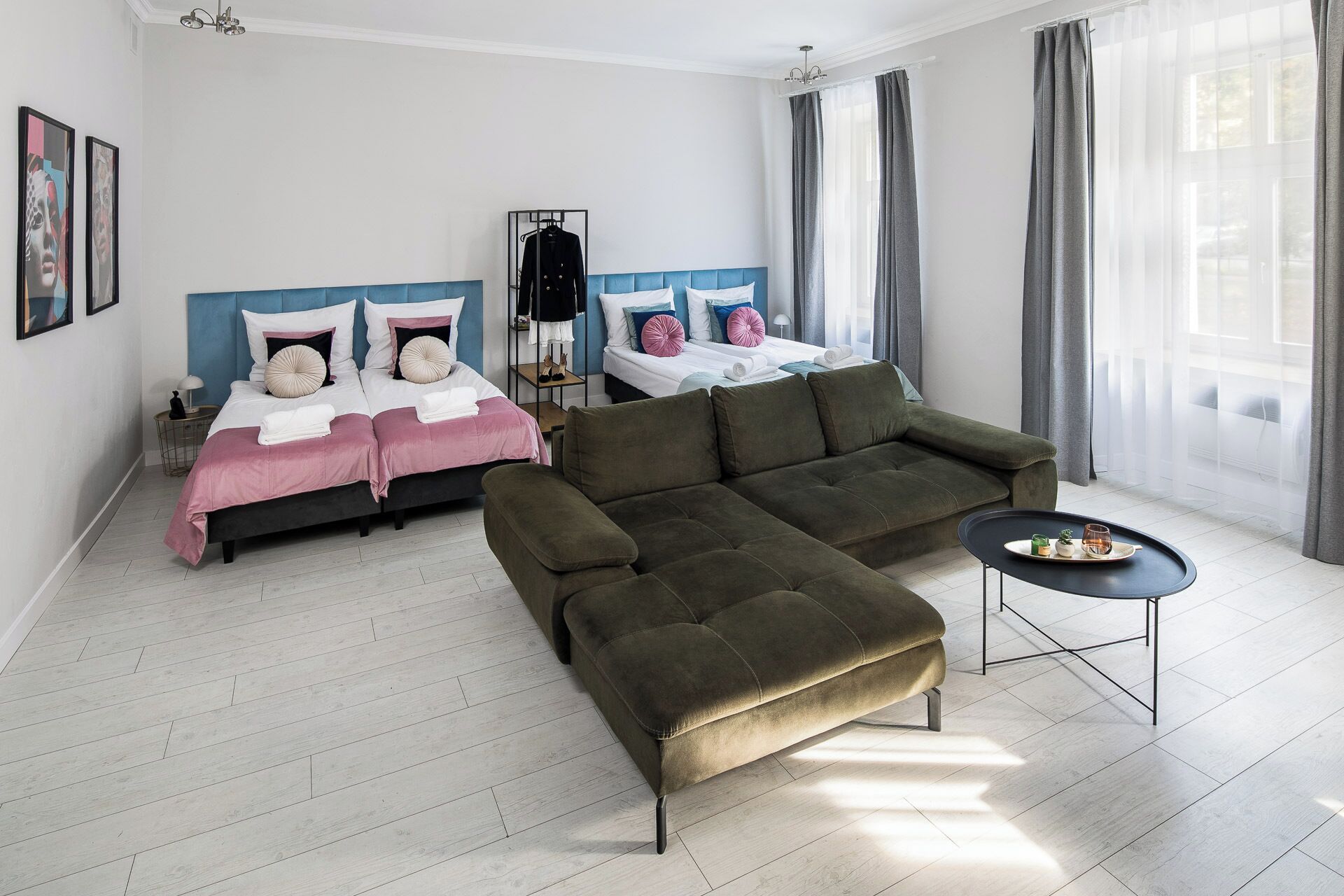 A modern hotel room with two beds, a green sofa, a small table, and a large window with curtains.