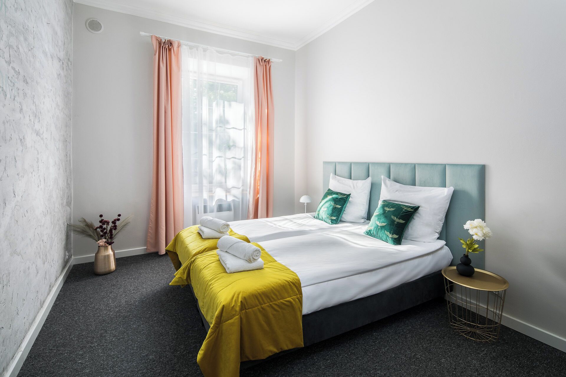 Habitación de hotel con una cama, dos mesitas de noche, un ventanal con cortinas y un jarrón con flores.