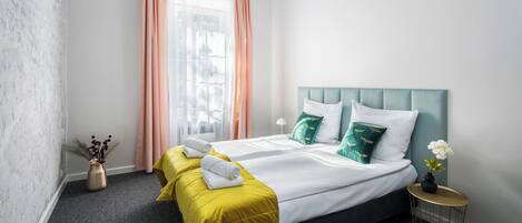 Habitación de hotel con una cama, dos mesitas de noche, un ventanal con cortinas y un jarrón con flores.