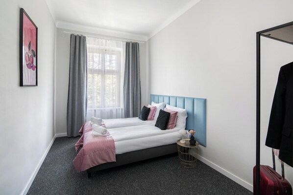 A hotel room with two beds, a large window with sheer curtains, a blue headboard, a round gold side table, and a red suitcase. - GA Apartments (Kraków)