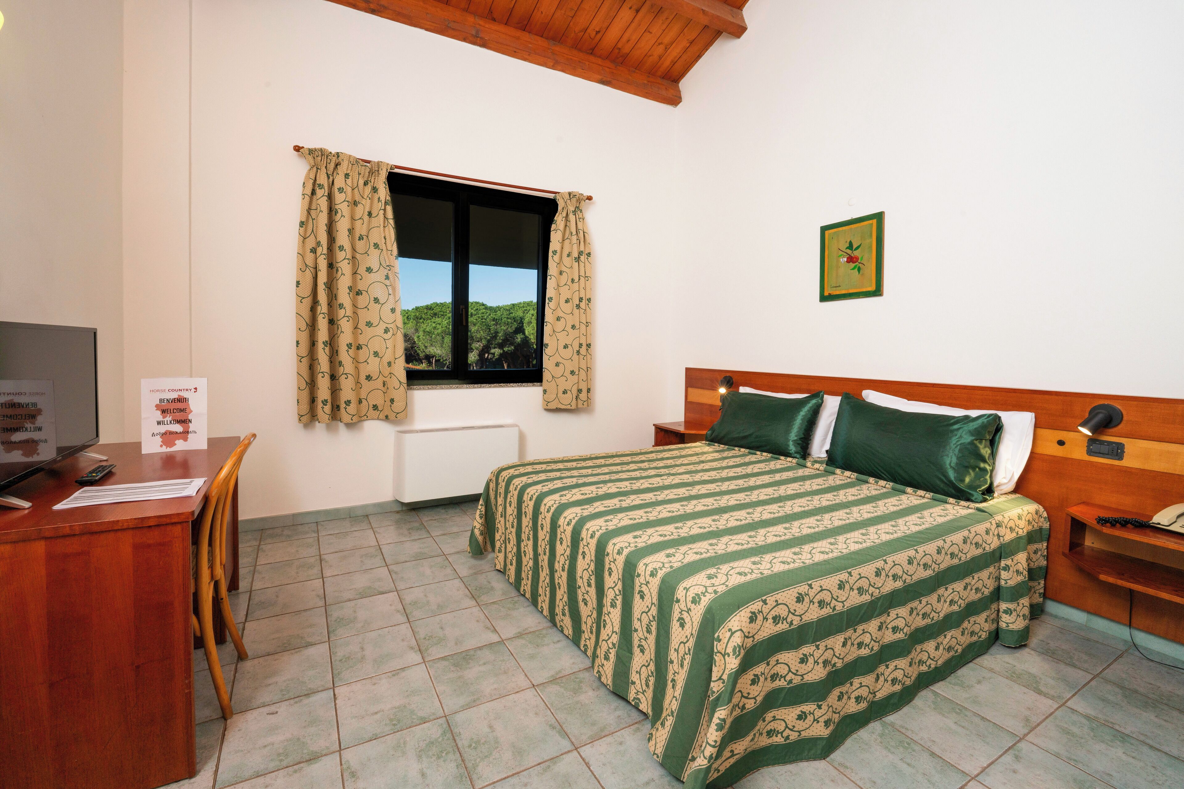 A hotel room with a bed, a desk, a chair, a window with curtains, a bedside table, a nightstand, and a framed picture on the wall.