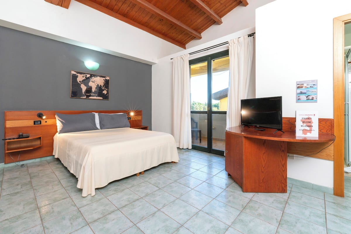 A hotel room with a bed, a desk with a TV, a world map on the wall, and a balcony with curtains.