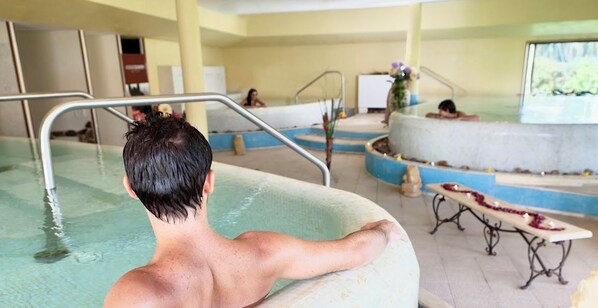 Zona de piscina con una persona relajándose en un jacuzzi, otra persona al fondo y un banco con flores.