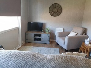 A living room with a television on a wooden stand, a grey armchair, a round wall clock, and a radiator. - Lovely Lodge Next To The Mansion House Llansteffan (Carmarthen)