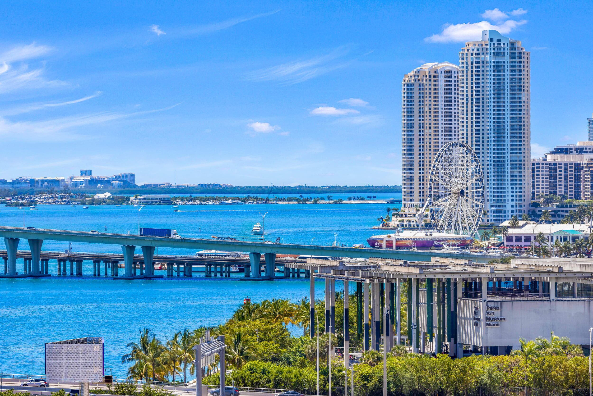 A coastal cityscape with a prominent Ferris wheel, a bridge, and high-rise buildings.