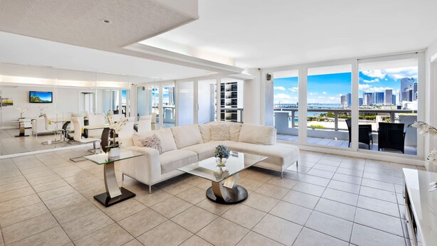 Uma sala de estar ampla com um sofá branco grande, uma mesa de centro de vidro e vista para o horizonte da cidade através de grandes janelas.