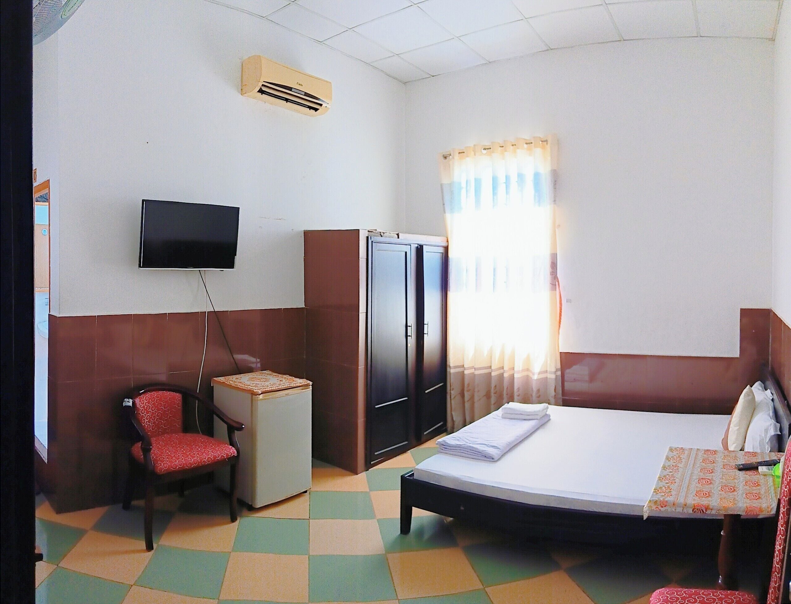 A hotel room with a bed, a chair, a small table, a TV, a refrigerator, and a window with curtains.