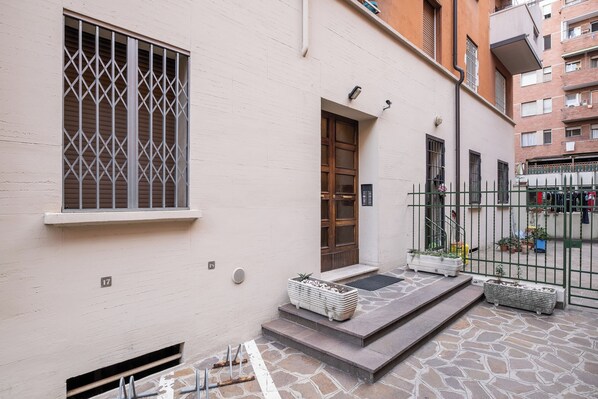 A building with a white exterior, a wooden door, and a window with metal bars. There are steps leading to the door, and potted plants are placed on the steps. - Porta Mascarella Apartment by Wonderful Italy (Bologna)