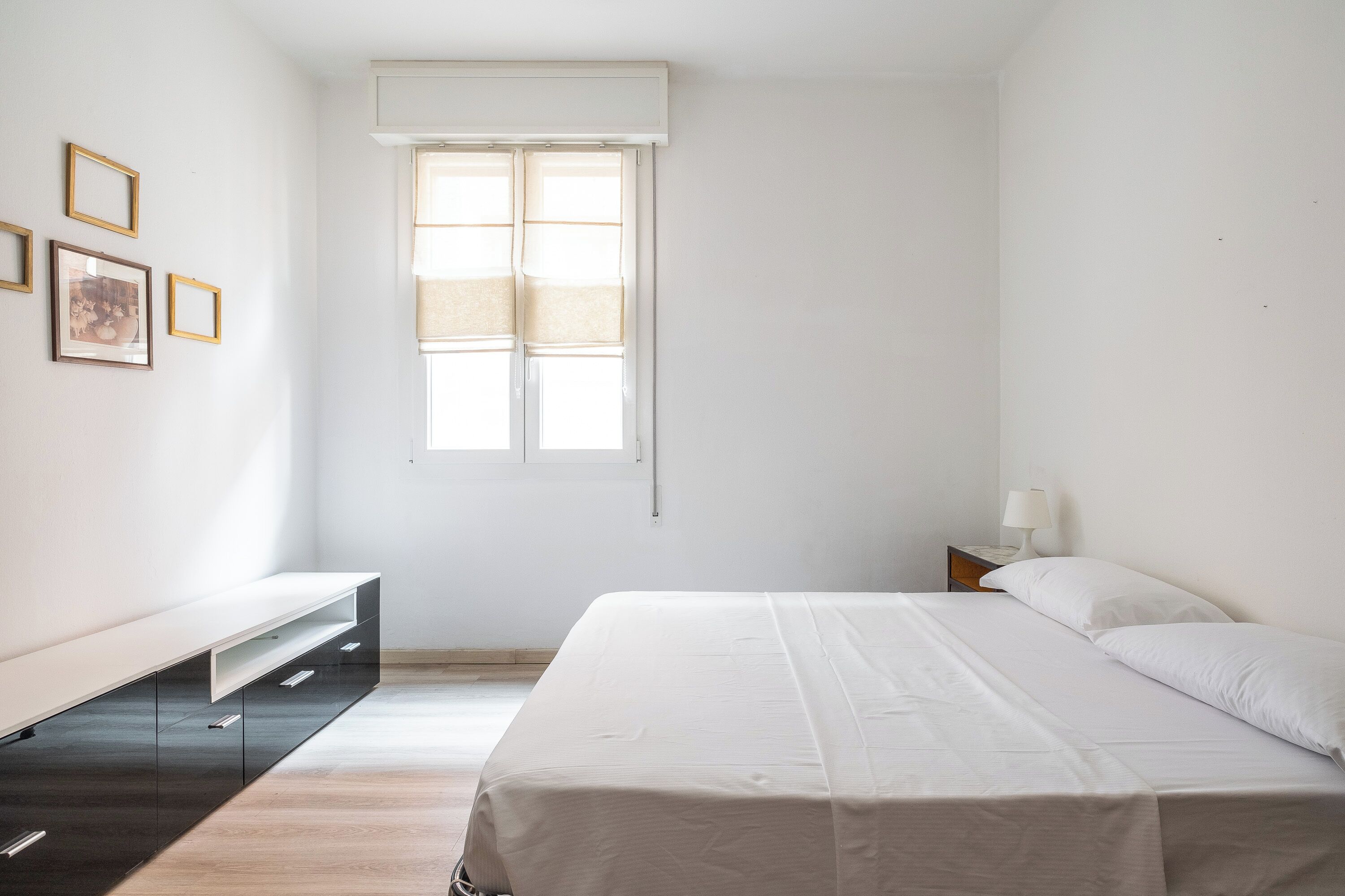 A minimalist bedroom with a bed, a bedside table with a lamp, a bench, and a window with blinds.