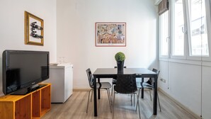Ett modernt vardagsrum med en platt-TV på ett träställ, ett matbord med stolar, ett litet kylskåp och ett inramat konstverk på väggen.